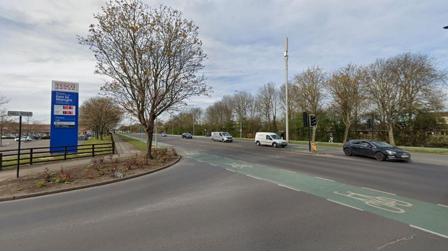 Cars travelling down a two-lane road. On the left is a Tesco petrol board showing and next to it is a road sing reading "Hall Road".