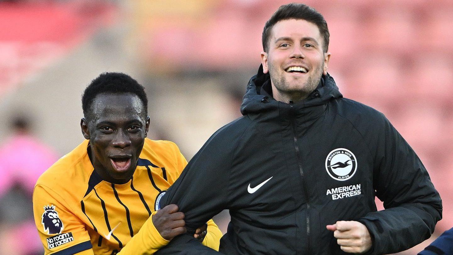 Yankuba Minteh, wearing a yellow Brighton shirt with narrow black stripes on the front, grabs the right arm of Fabian Hurzeler, who is wearing a black Brighton-branded training jacket. Minteh is opening his mouth to speak and Hurzeler is smiling as both men look towards the camera