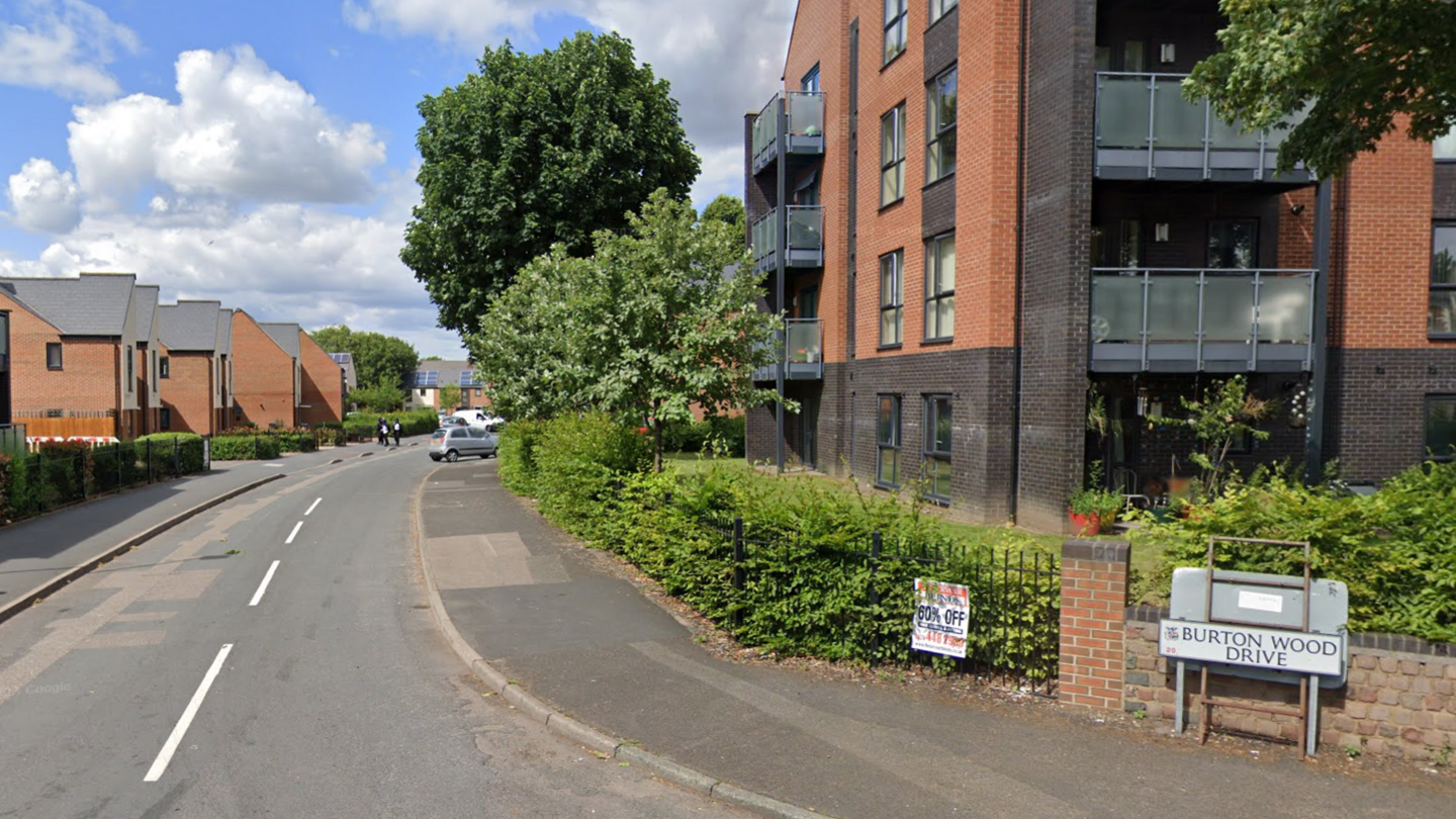 A residential street with houses and blocks of flats, with a road sign saying 'Burton Wood Drive'