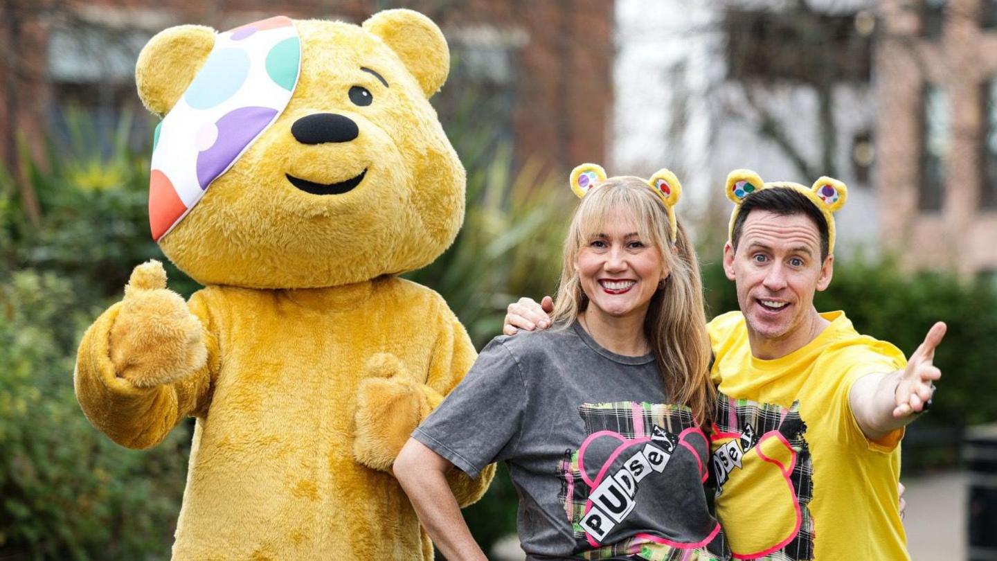 Pudsey bear, a yellow bear with a spotty eyepatch alongside Cate Conway, who has light hair. She is wearing yellow bear ears and a grey top, she is beside Conor Phillips who has dark hair. He is wearing yellow bear ears a yellow top.