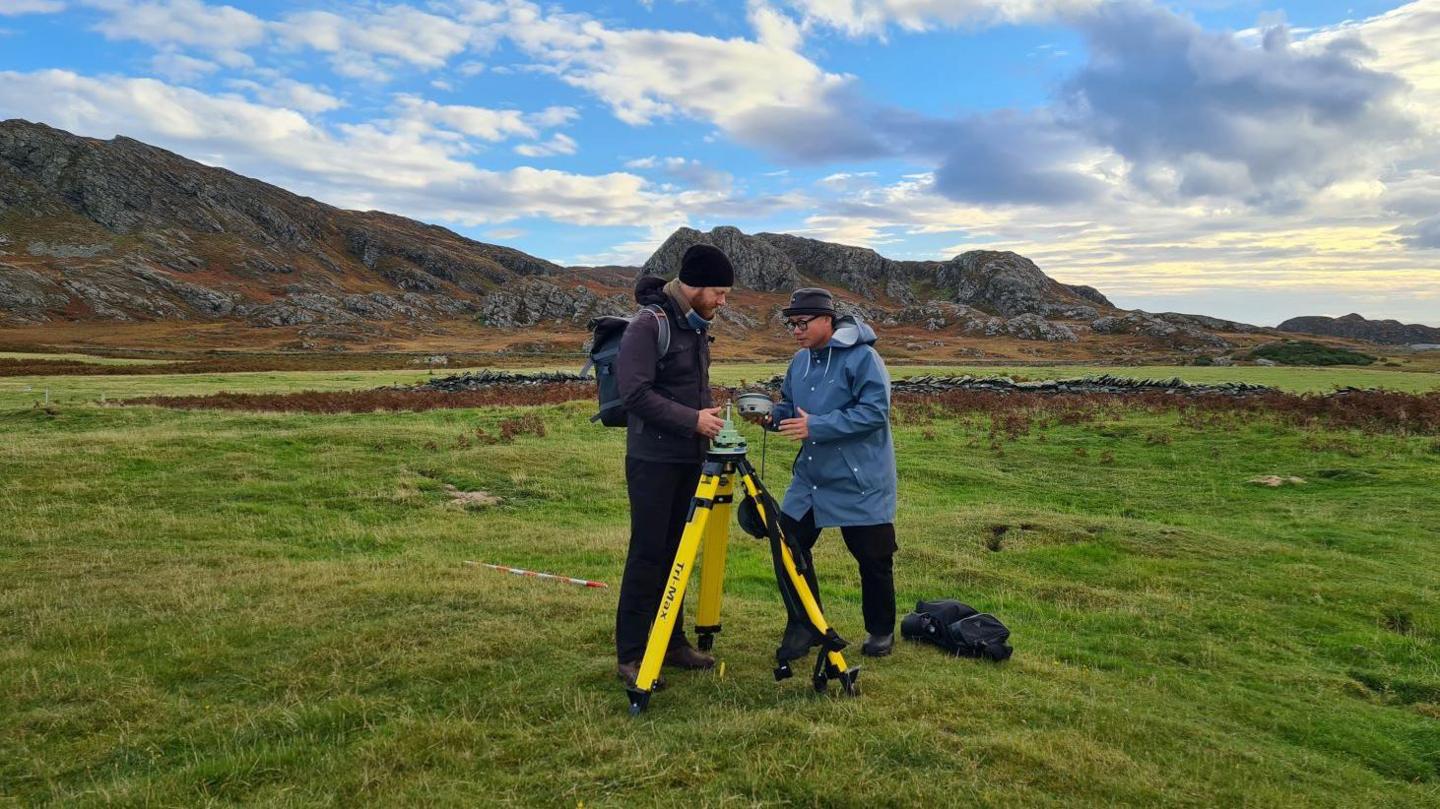 Two German project members in a grassy field setting up GPS equipment for fieldwork and conducting survey using ground penetrating radar to discover underground remains. There are rocky hills in the distance.