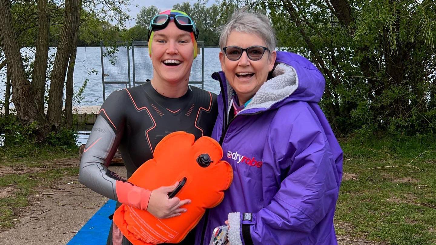 The pair near to a lake, with the daughter in a wetsuit and goggles and holding a float, and the mother in a purple dryrobe