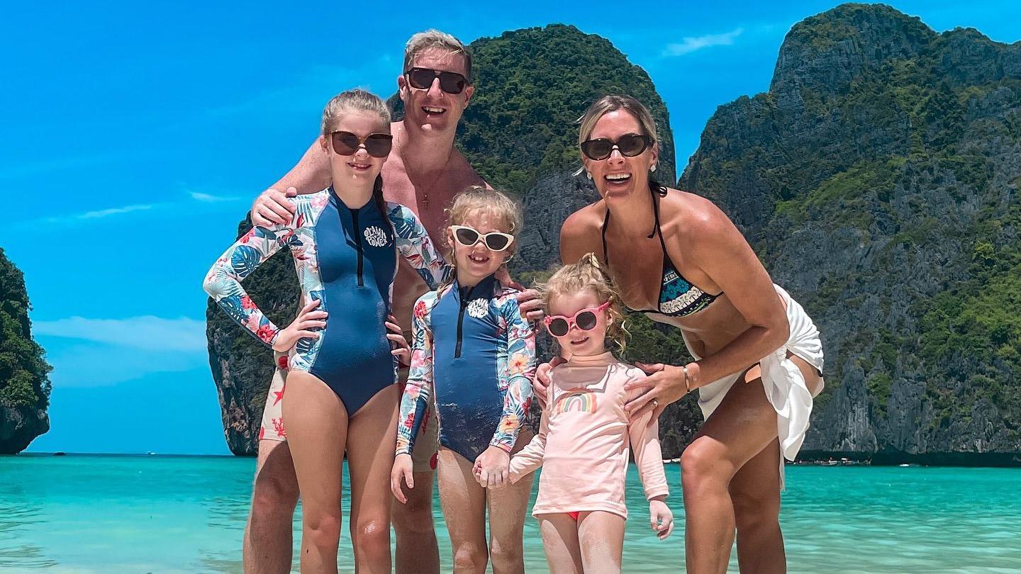 The Hutchinson family - Chris and Tamira and their three daughters - standing in front of turquoise blue sea with grassy large rocks in the background.