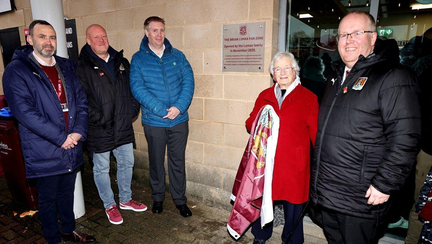 Four men and one woman - who is holding a Northampton Town flag - pose in front of a plaque at the Brian Lomax Fan Zone.