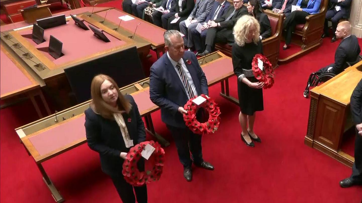 Speaker Edwin Poots holds a poppy wreath as he leads an Armistice Day ceremony at a room in Stormont. He is flanked by two officials also holding poppy wreaths while several people are gathered on benches behind them