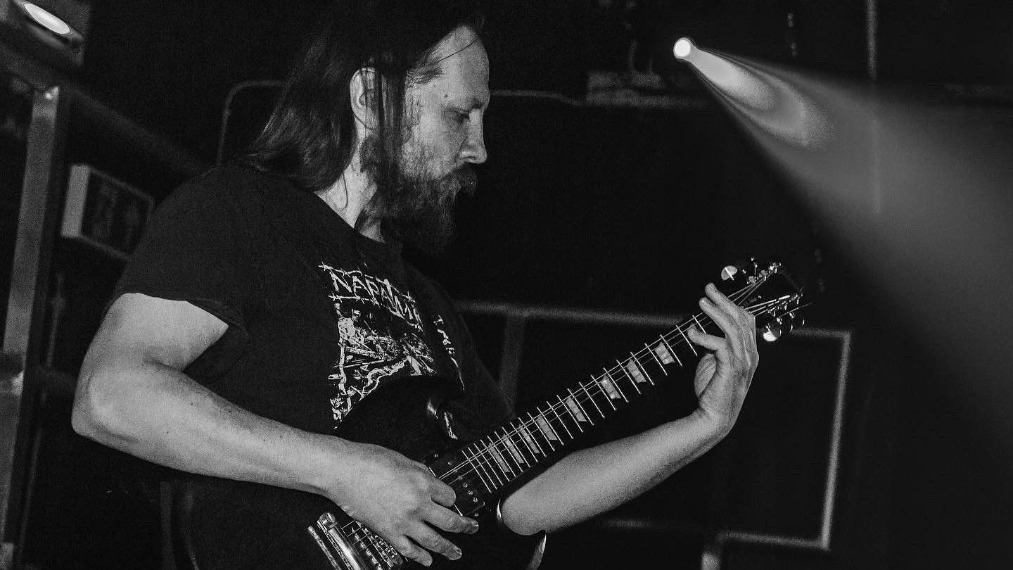 A black and white image of Luke who's playing the electric guitar on stage. He has a beard and long hair, and is wearing a black T-shirt with white writing on it