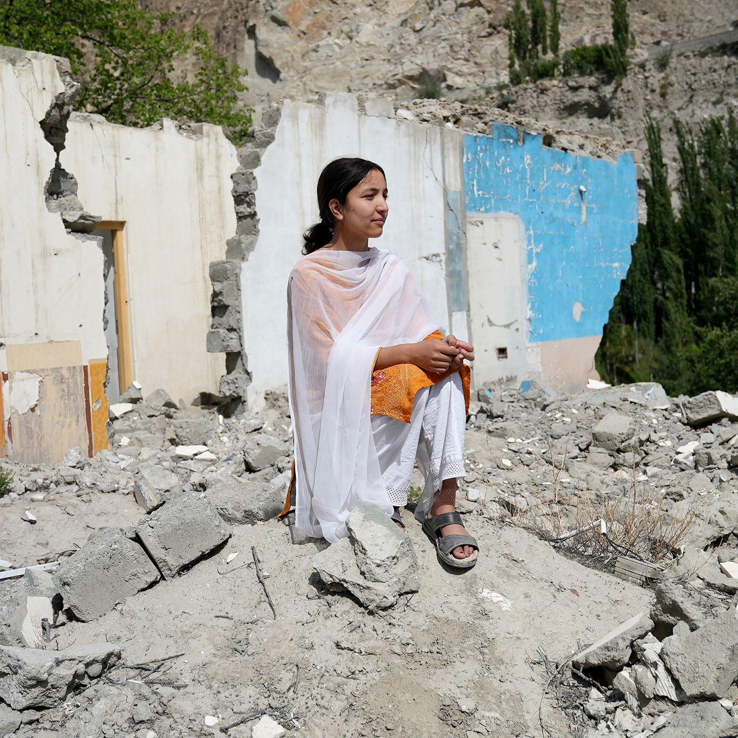 Komal Sher looks out to the right of the camera while wearing an orange top, snadals and white shawl. She is standing on the rubble of buildings in her village of Hassanabad, with damaged walls in the background.