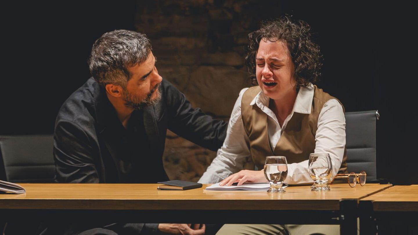 A woman sitting crying at a table with a man crouching down beside her with his arm on her back. There are glasses containing water on the table as well as a notebook, papers and the woman's glasses. The man has dark hair and a beard and is wearing a dark blazer over a dark shirt. The woman has short, curly hair and is dressed in a white shirt and a tan waistcoat and trousers. Her face is scrunched up showing her distress. 
