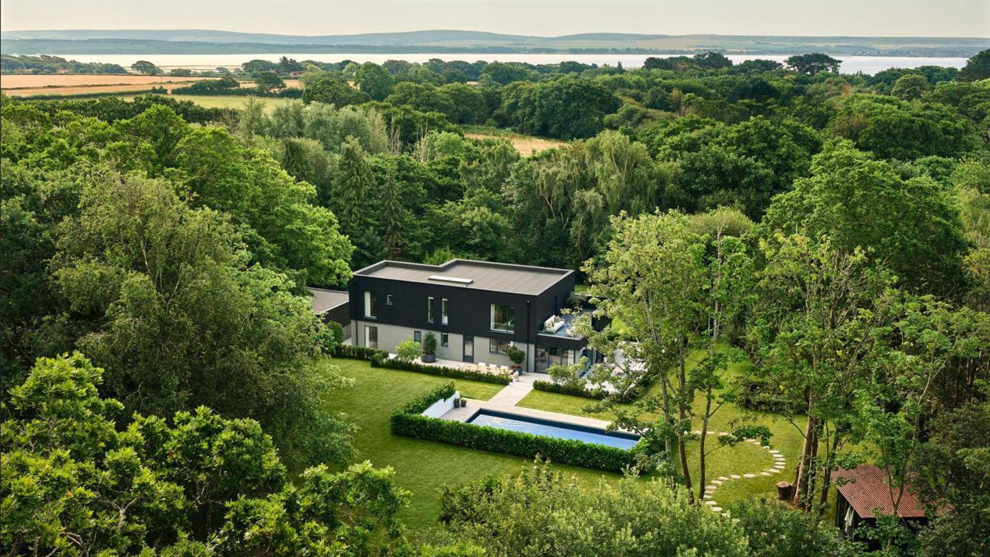 A drone shot of the house, the sea is visible in the background. Trees encircle the property and a swimming pool bordered with a hedge is next to the house.