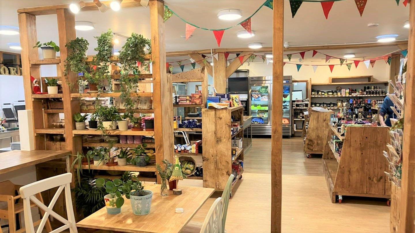 A view inside the café towards the shop. There are large wooden shelving units which have been decorated with potted plants. There is colourful fabric bunting hanging from the wooden ceiling rafters. In the background there is the shop, with fridges and shelves full of local and organic produce.