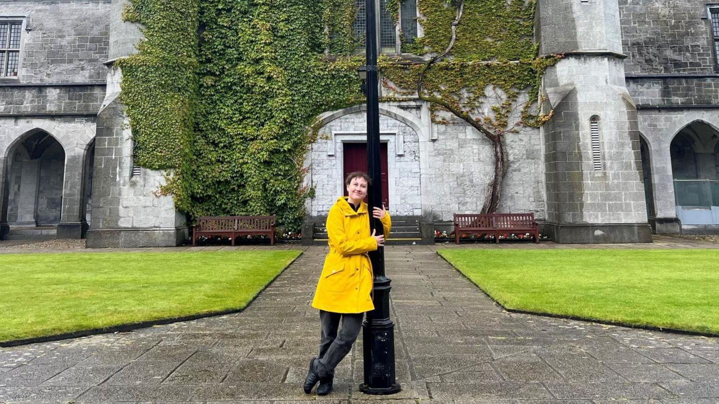 Gabriella Moro stands holding a lamppost in a bright yellow trench coat in the square at the University of Galway.