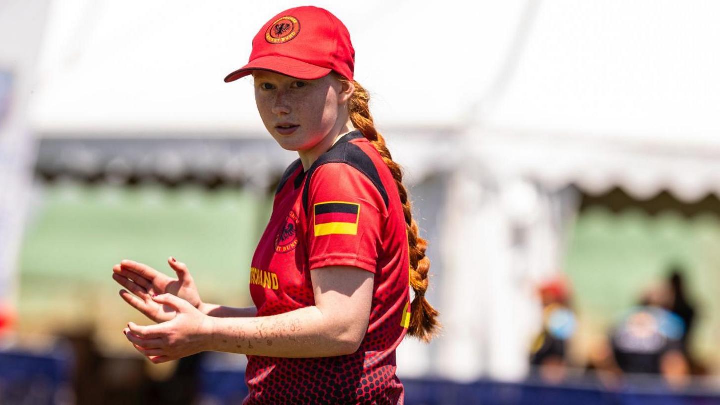 A young girl is wearing a red cricket kit showing the Germany flag. She wears a red cap over her ginger hair, which is in a braid. She's clapping while looking away from the camera, seemingly at her team mates.