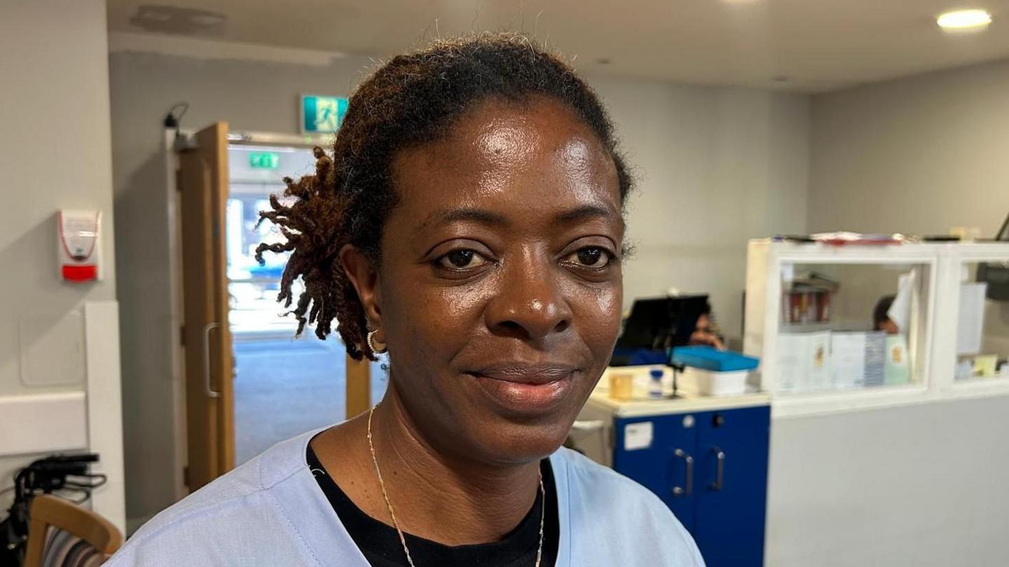 Folake looking to the right of the camera. She is wearing her blue care home scrubs with a thin gold chain and a black layer underneath. Her dark hair is tied back. A care home reception desk can be seen to the right hand side, with an open door behind her.