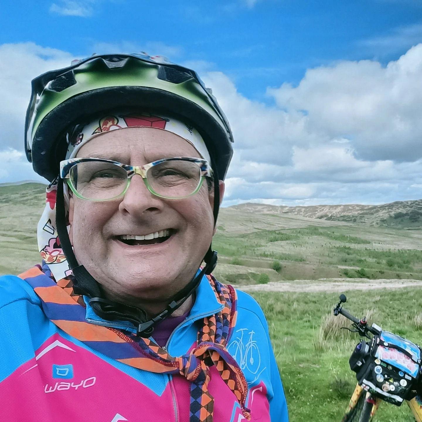 Timmy Mallett in a colourful cycling top with a bike helmet on and a orange and blue neckerchief. His bike is resting in the background on a sprawling Dumfries and Galloway country scene.