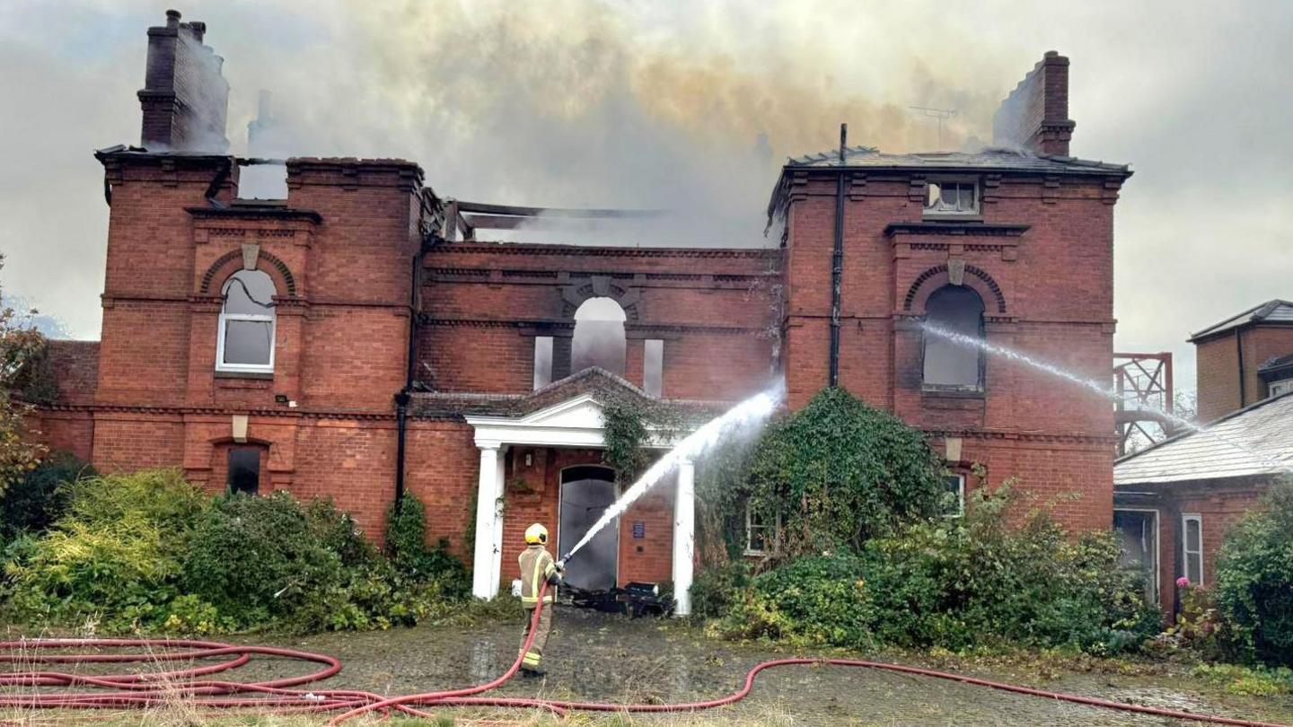 A firefighter hosing down a large double fronted property with water. The roof of the building has been gutted and many of the windows have also blown out as a result of a fire. There are no visible flames, but dense smoke is still rising from the structure.