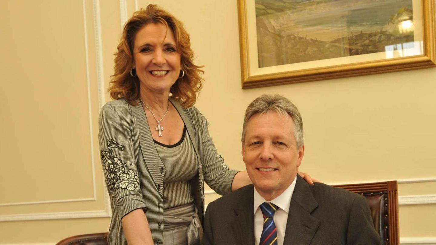 Iris and Peter Robinson smiling in an office. Iris has long red hair and is wearing a black and white top with a cardigan. Peter has short grey hair and is wearing a grey suit with a white shirt and a blue, yellow, dark blue and red striped tie.