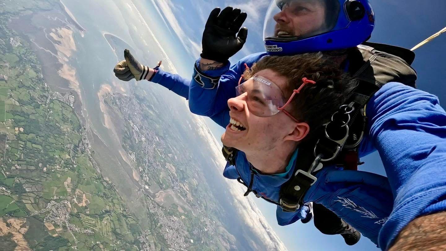 Charlie is in the air strapped to an instructor. They are wearing a blue diving suit. You can see the land from up above.