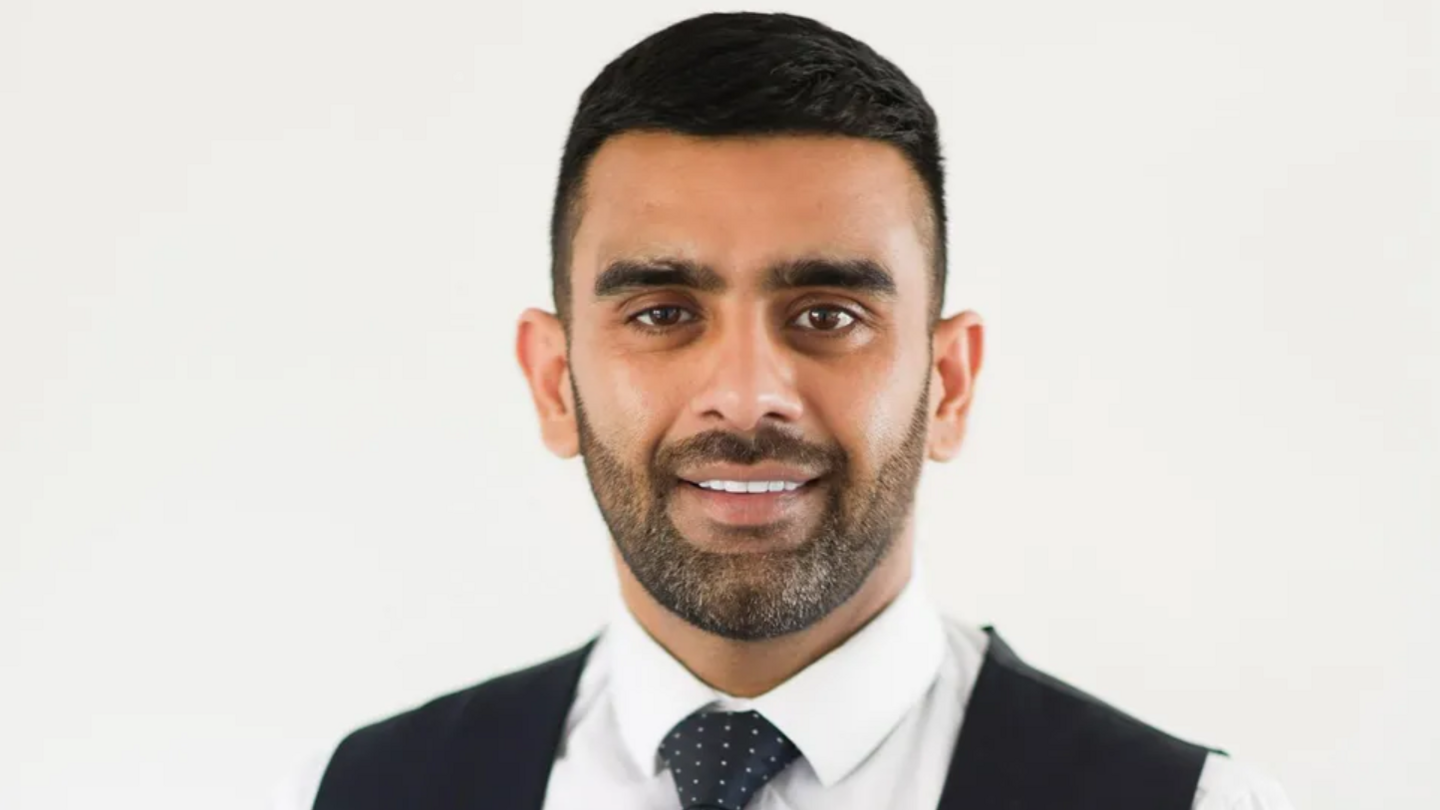 A close-up picture of Akhmed Yakoob, smiling at the camera in front of a white background. He wears a white shirt, navy tie, and a dark jacket.