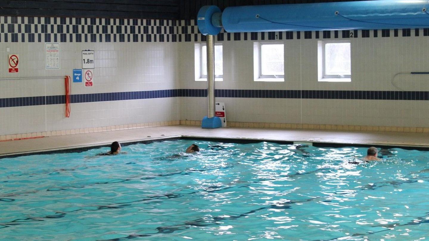 An indoor six-lane swimming pool, 25m (82ft) long with some swimmers doing lengths. The walls have black and white chequered tiles and there are beige coloured non-slip tiles on the walkway around the pool.