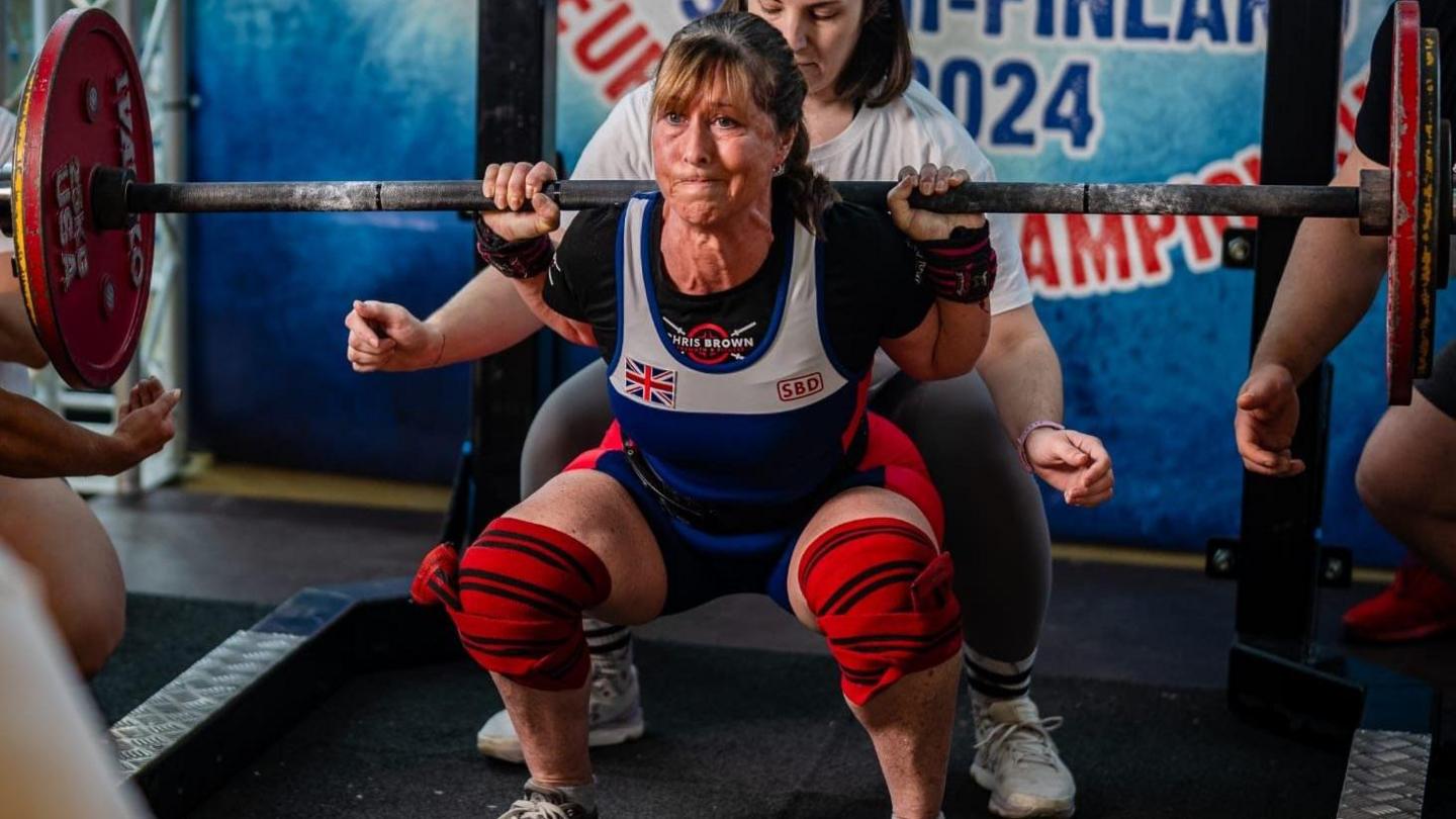 Louise Clark wearing a Great Britain competing tank top and shorts, with red knee protection bands as she lifts in th powerlifting competition in Finland.