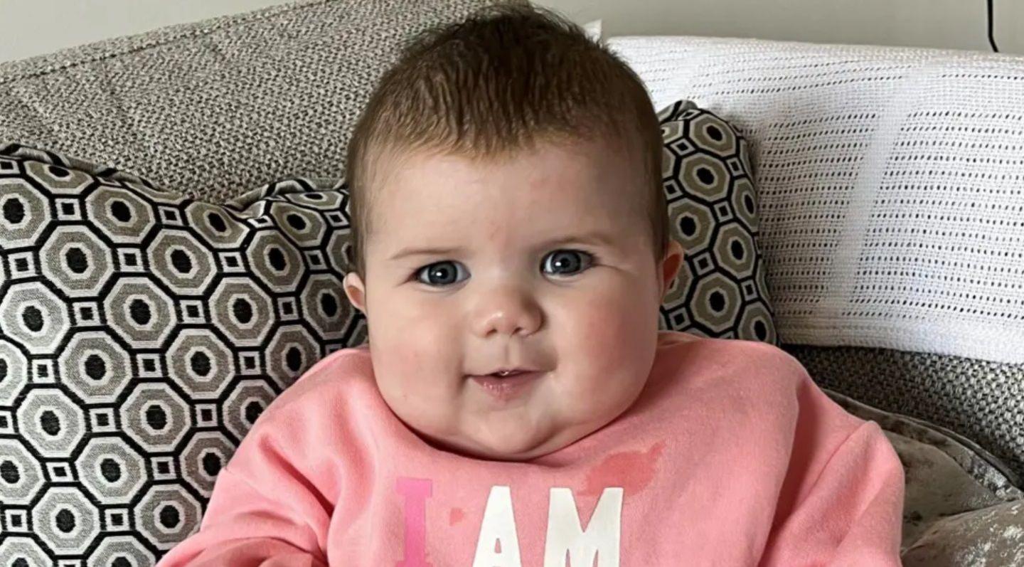 Baby Mabli looks at the camera. She has short dark hair and is wearing a pink sweatshirt. She is resting against a pillow with black and grey geometric designs on, and is sitting on a grey sofa. 