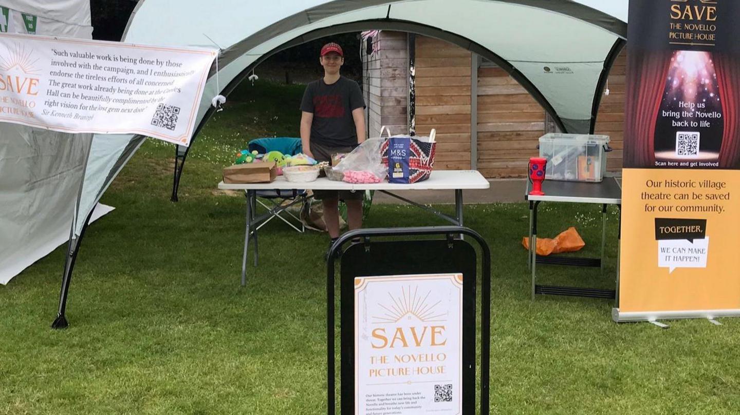 A stand at a fair with signs and posters about the Novello Theatre campaign. A person is standing under a gazebo behind a table laid out with bags and other items.