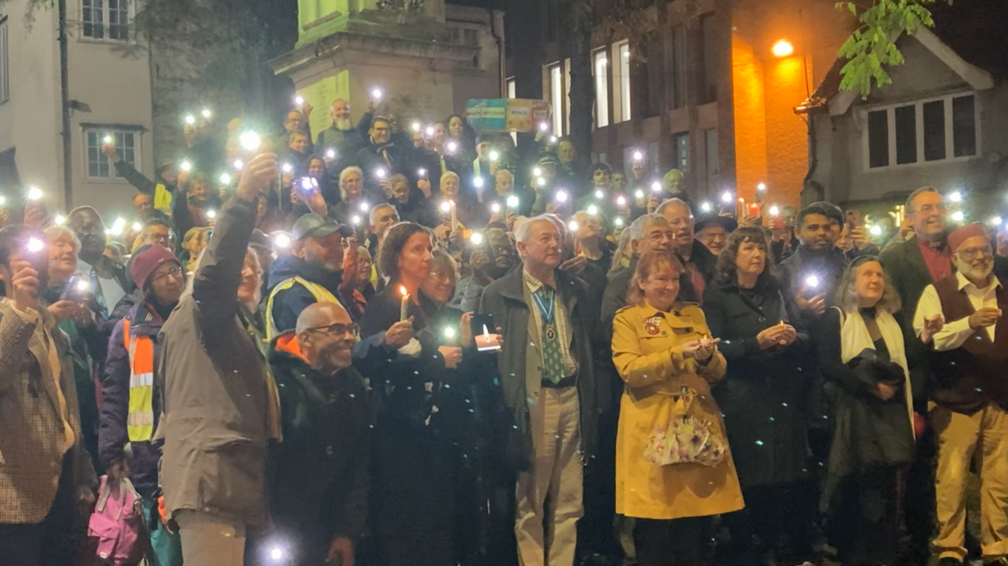 About 50 people standing together on a street. They are holding their phones in the air, and their phones have their torches turned on