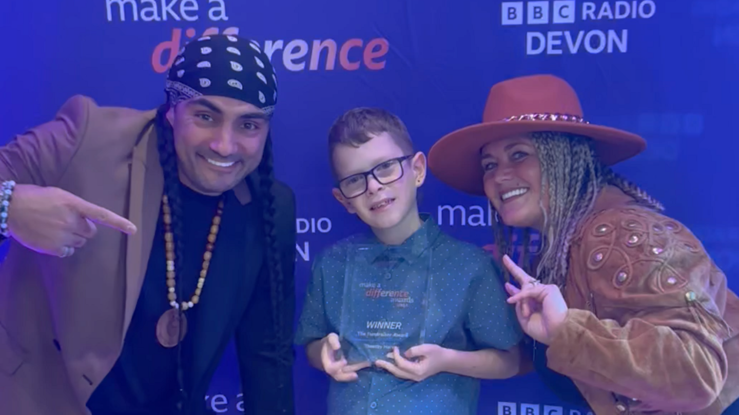 10-year-old Tim holding his glass Make a Difference award. He is wearing glasses and a blue shirt with a white polka dots He is stood against a purple background with the words 'Make a Difference' and 'BBC Radio Devon' along with the judge Toby Gorniak (left) and Jo Gorniak (right) either side. 