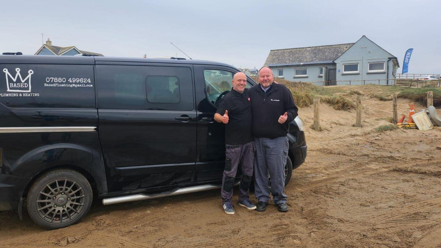 Plumber Daniel Terry, left, stood next to another man and his company van, which is on a beach.