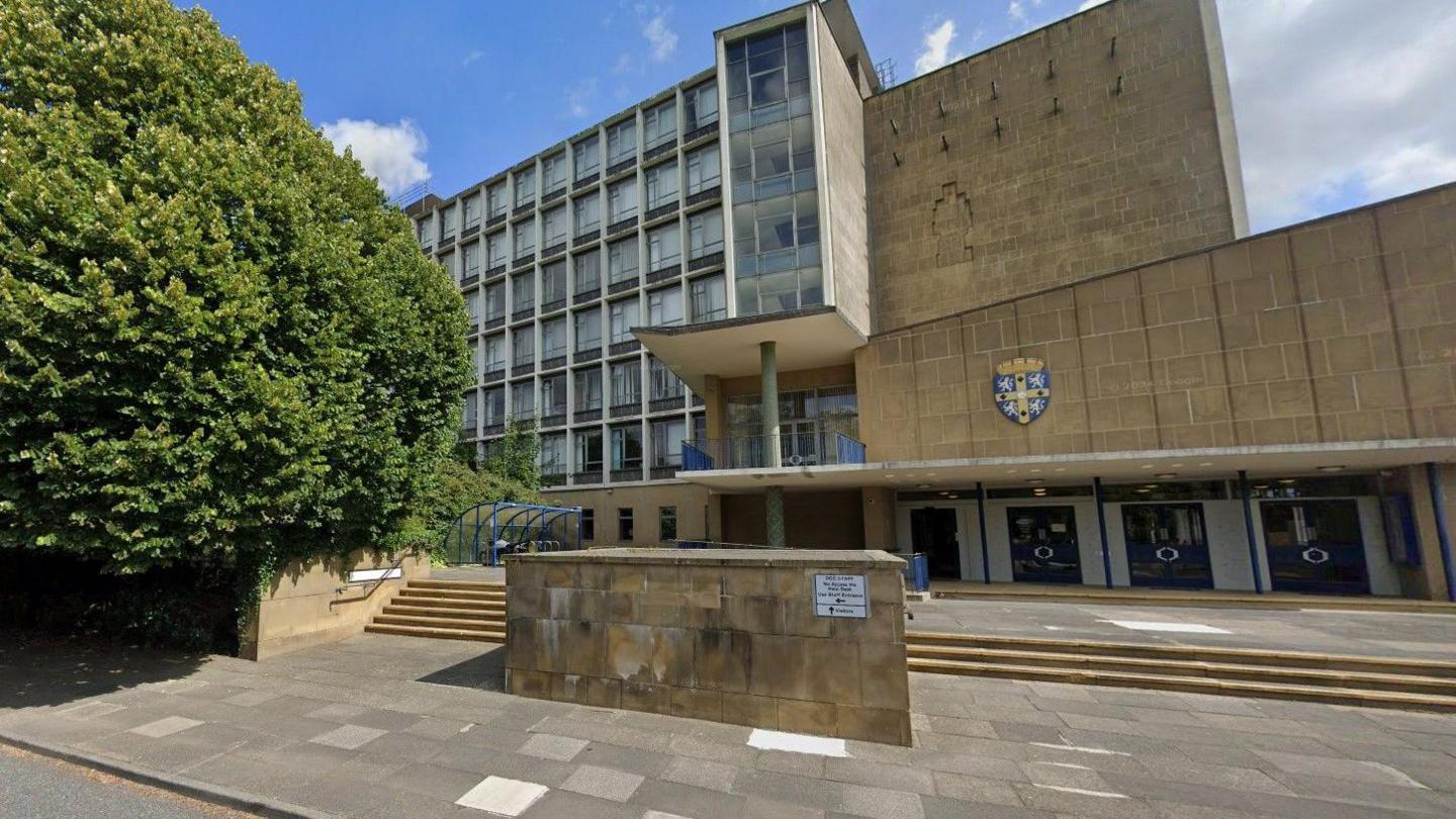 Durham County Hall. It is a seven-storey building with a lot of windows and small steps leading to the entrance. There is a blue, white and yellow crest above the glass entrance doors to the right. There is a large tree and a bike shed on the left side in front of the building.