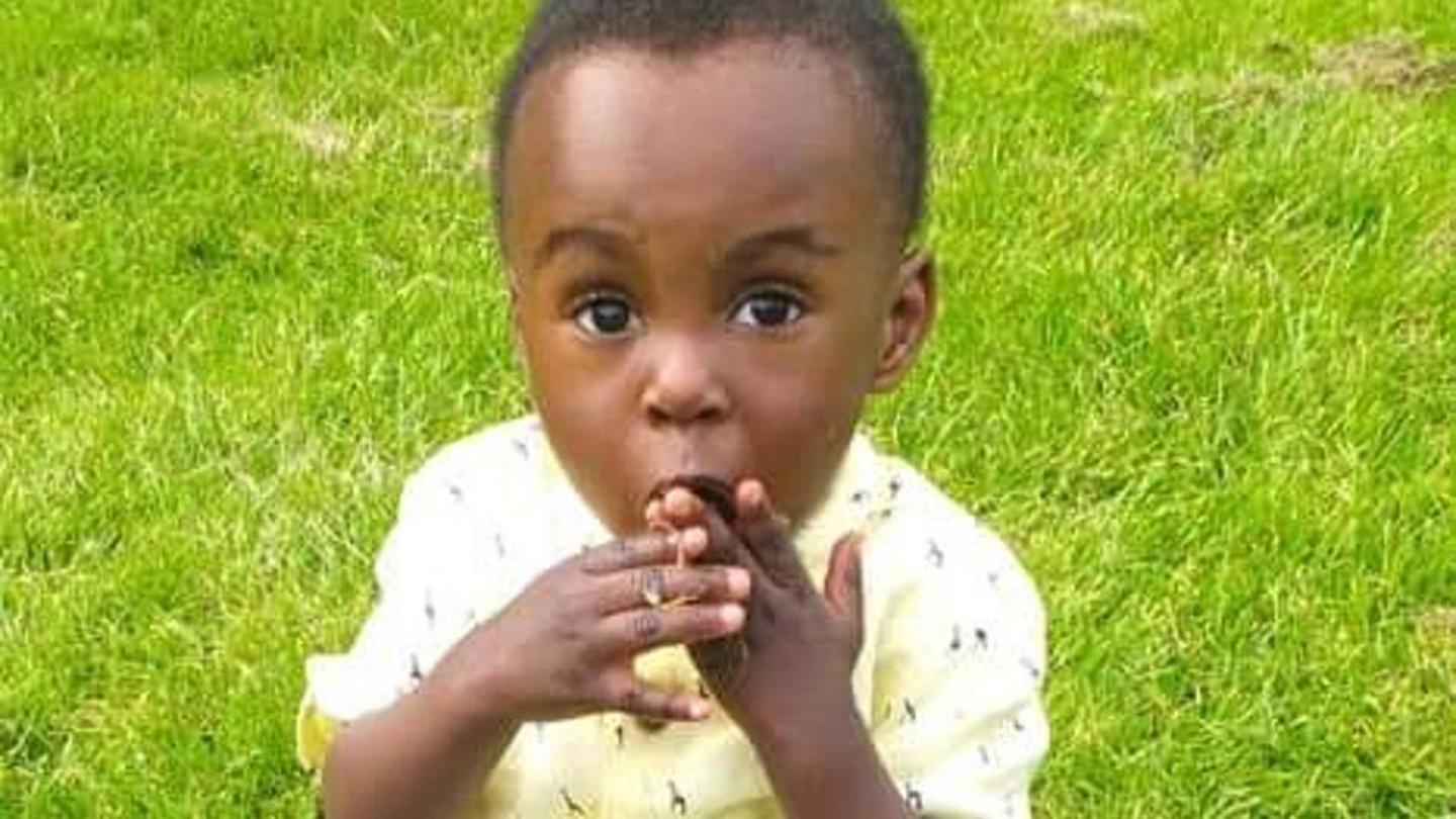 Awaab, a young boy, looks at the camera as he sits on grass. He has his hands in his mouth and is wearing a yellow shirt.