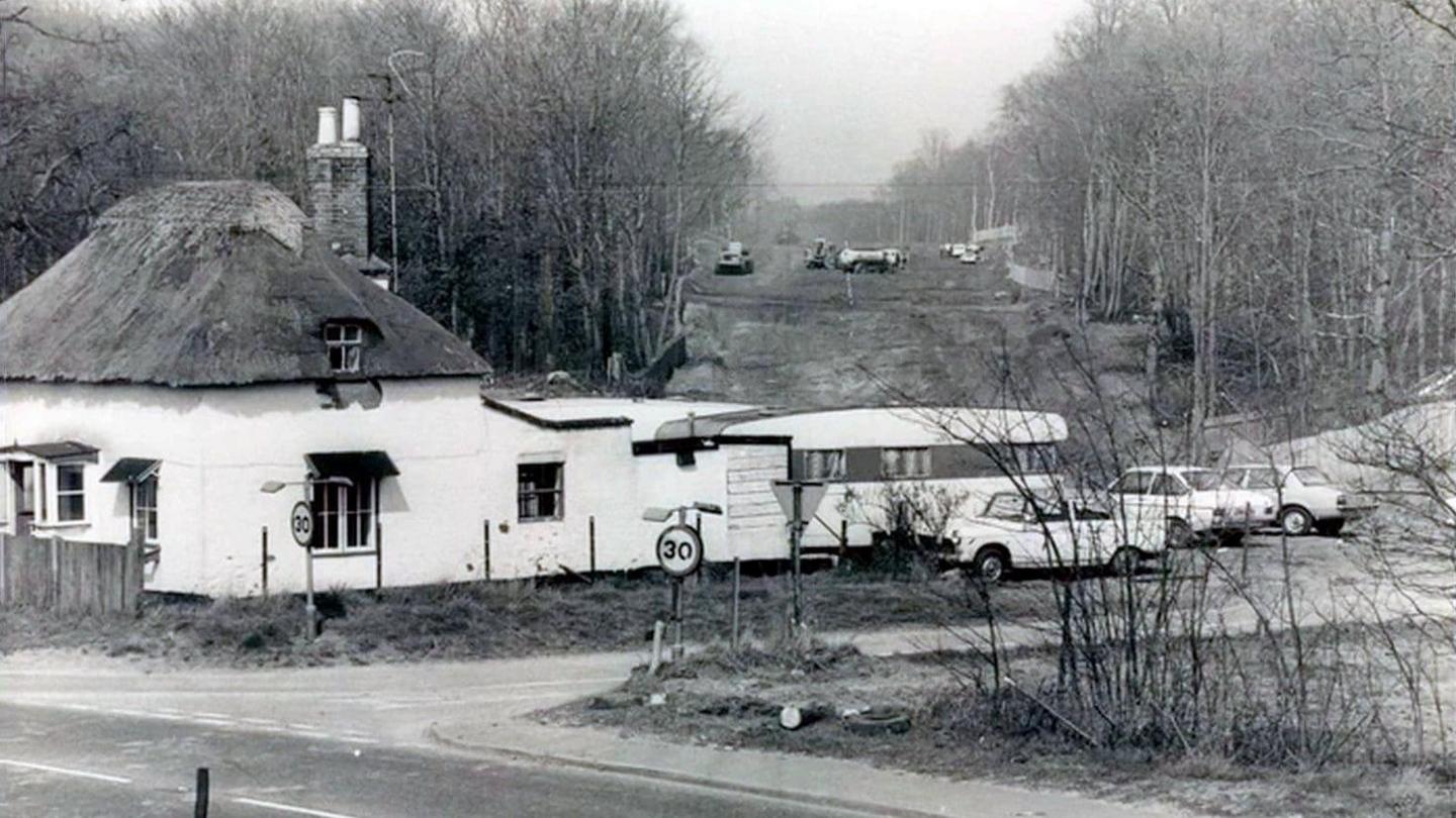 A black and white photo of a house with a caravan and cars parked to the back of it, with construction going on in the background. 