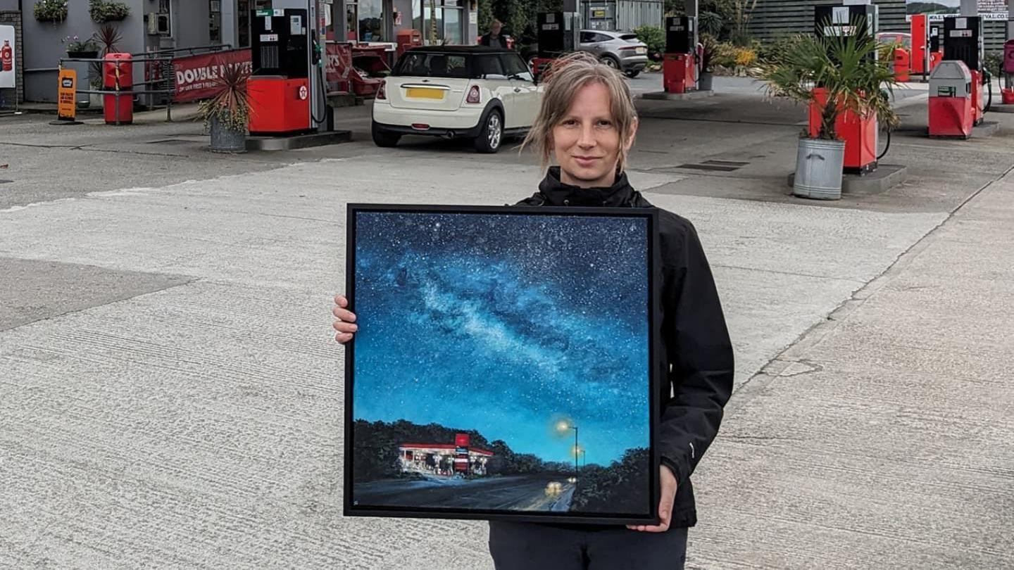 Diane Griffiths stands on the forecourt of the Texaco petrol station holding her artwork. Ms Griffiths is wearing a black jacket. She has brown hair. The painting is in a black frame. The painting shows a dark starry sky over a red and white petrol station. Street lamps are lit up and cars have their head lights on.