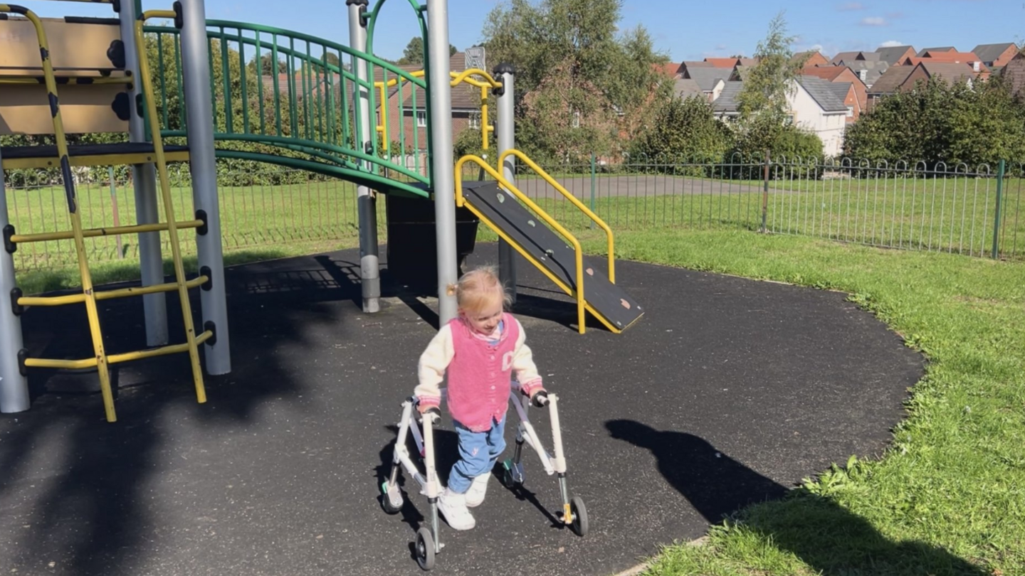 A three-year-old girl, Dakota, is in the playground using her frame by climbing equipment. There is a yellow climbing frame in the background on a hard surface with grass surrounding it further away. Dakota has blue trousers on and a pink top.