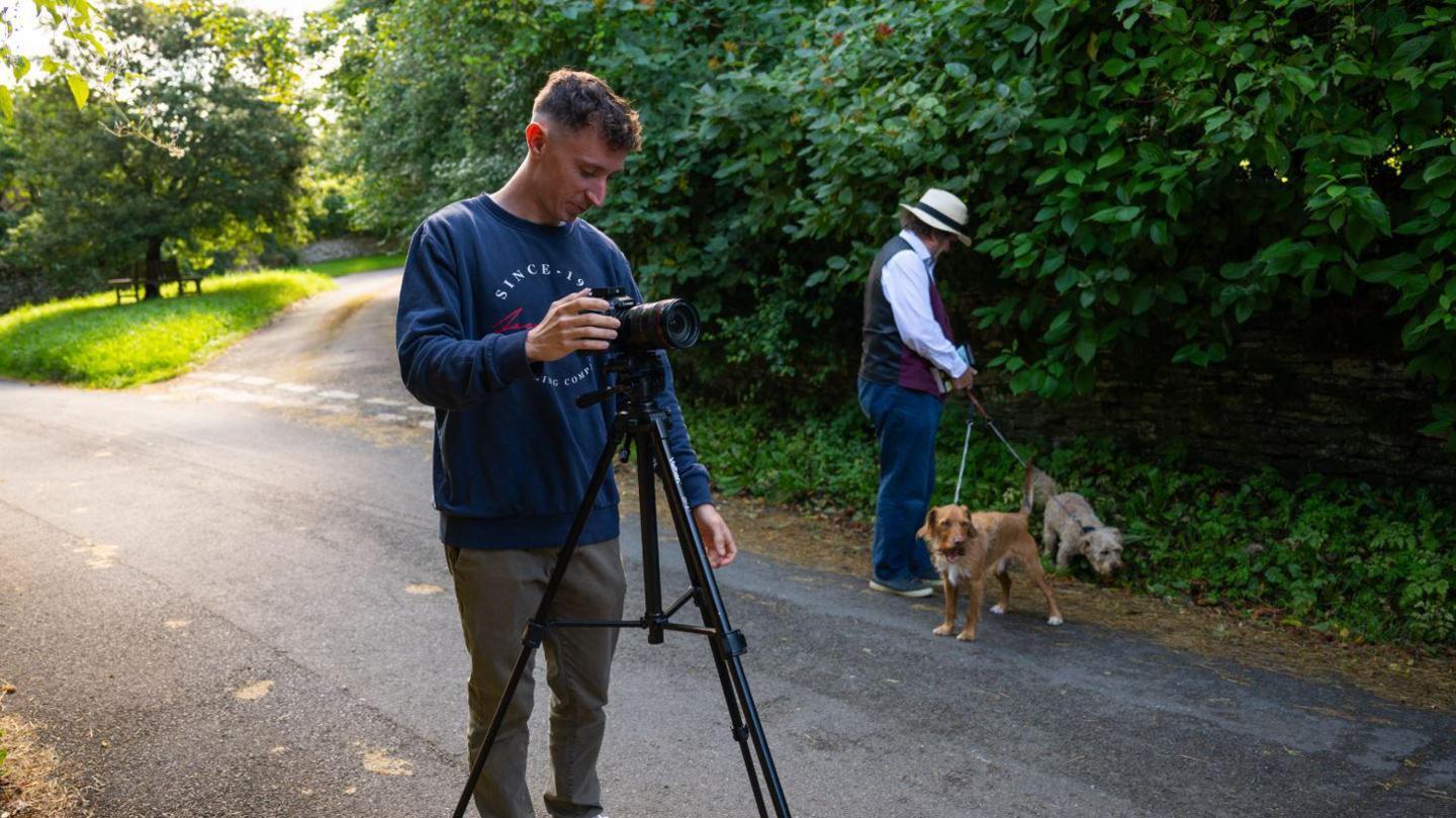 Robin Shuckburgh and Ross Arrowsmith filming on a countryside road. Mr Shuckburgh is looking after his dogs while Mr Arrowsmith is setting up the camera. It's a sunny day.