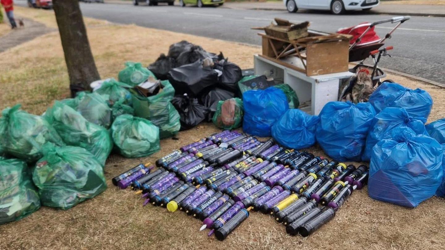 82 purple cylinder-shaped canisters on the grass. They are surrounded by green and blue plastic bags full of rubbish.