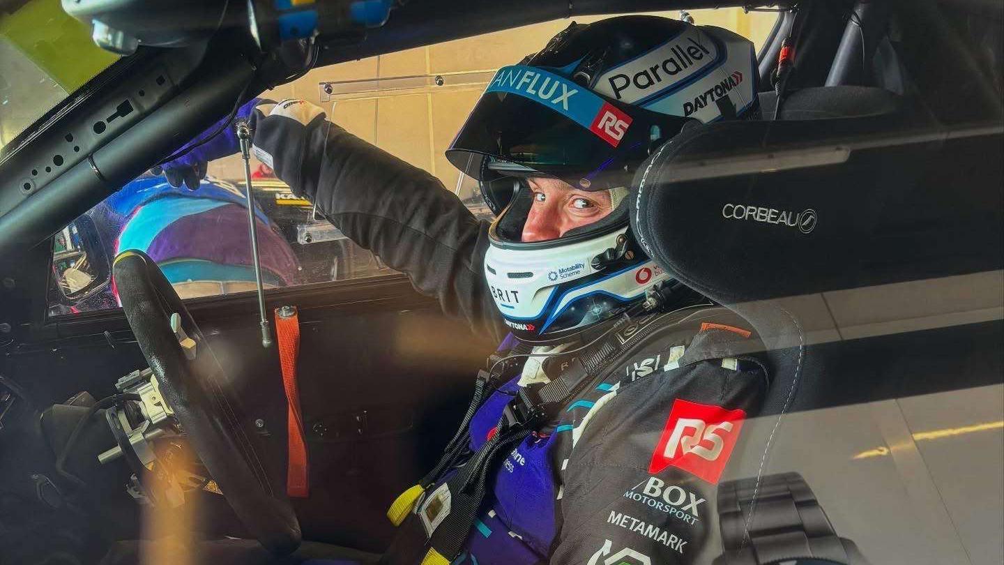 A race car driver is seated inside a race car, strapped in and wearing a helmet, looking toward the camera through the car window.