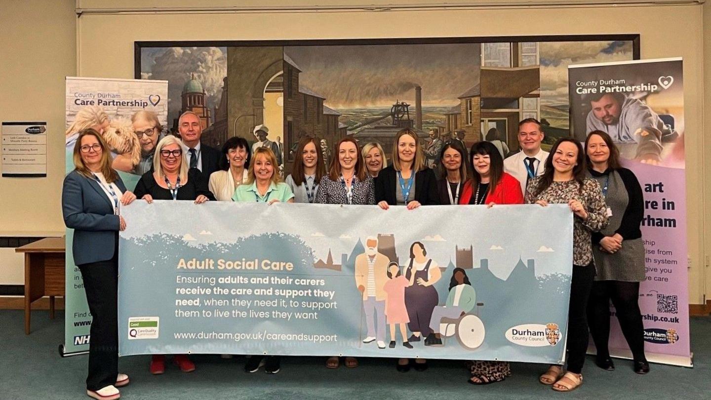 Group of 14 adults, mainly women, smiling and holding a banner for Durham County Council's adult social care service.