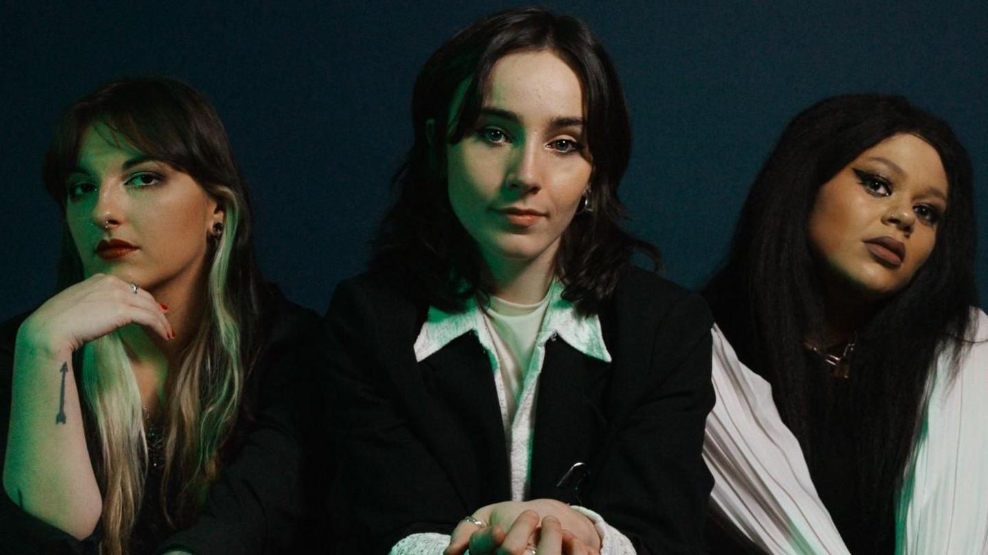 CHERYM , made up of L-R Alannagh Doherty, Emer McLaughlin and Hannah Richardson. The band are dressed in monochrome outfits and sit posing on a black sofa against a dark blue backdrop.