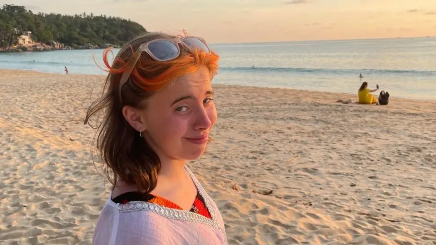 A young woman stands on a beach. She is smiling at the camera.