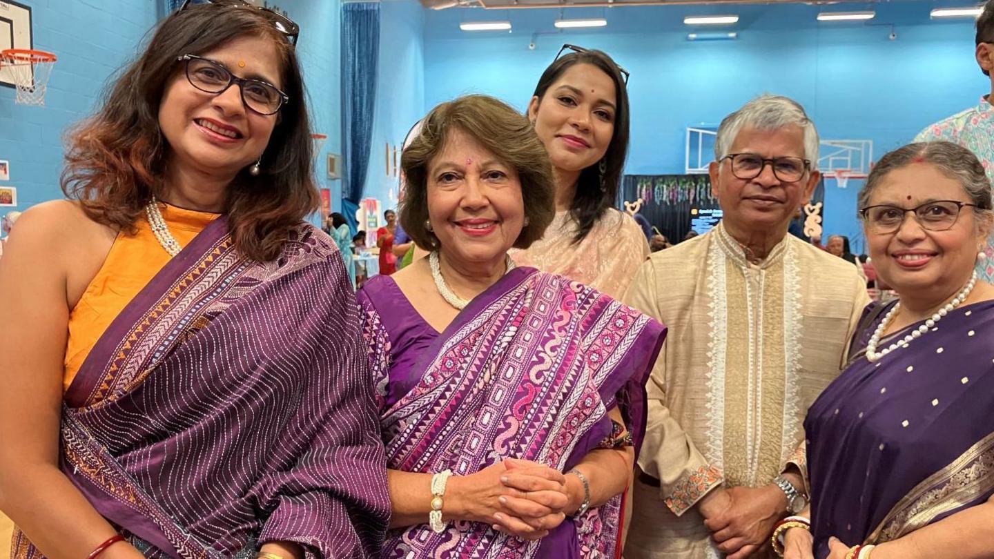Four women and one man wearing traditional Bengali outfits, stand smiling at the camera. They are inside a sport centre with a basketball hoop visible in the background