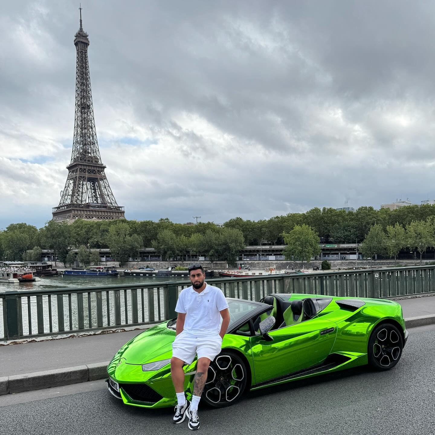 Gurvin Singh Dyal is wearing white shorts, t-shirt, sock and trainers as he looks ar the camera while leaning on the bonnet of a bright green metalic supercar. The car is parked next to a pavement on a bridge with part of the River Siene visible and the Eiffel Tower in the background. The sky has hints of blue but it mostly cloudy. 