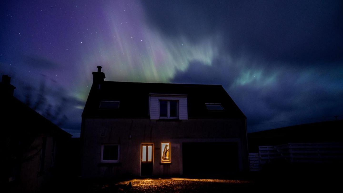 A house with lights on at the front door. Behind it the aurora appears in colours of green and purple.