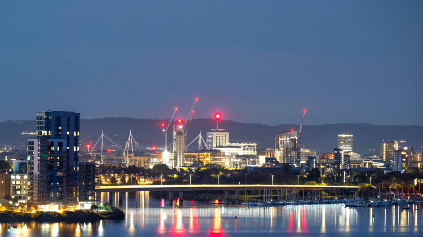 Cardiff skyline at night. There are high rise buildings and cranes in the background, with the Cardiff Bay flyover illuminated. The skyline reflects in the sea below. 
