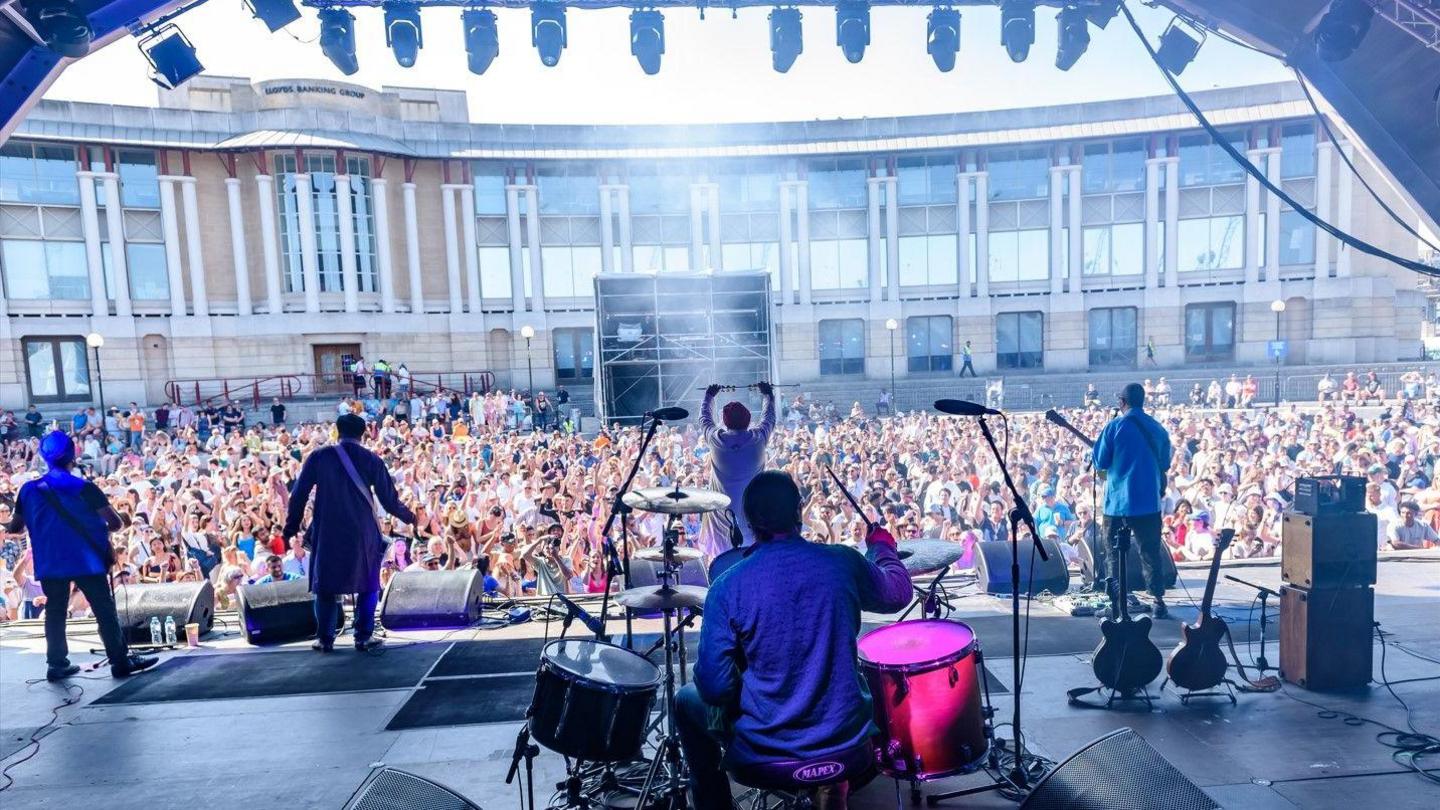 A view from the back of a stage looking at a five piece band  with their backs to the camera on a large stage facing a large audience