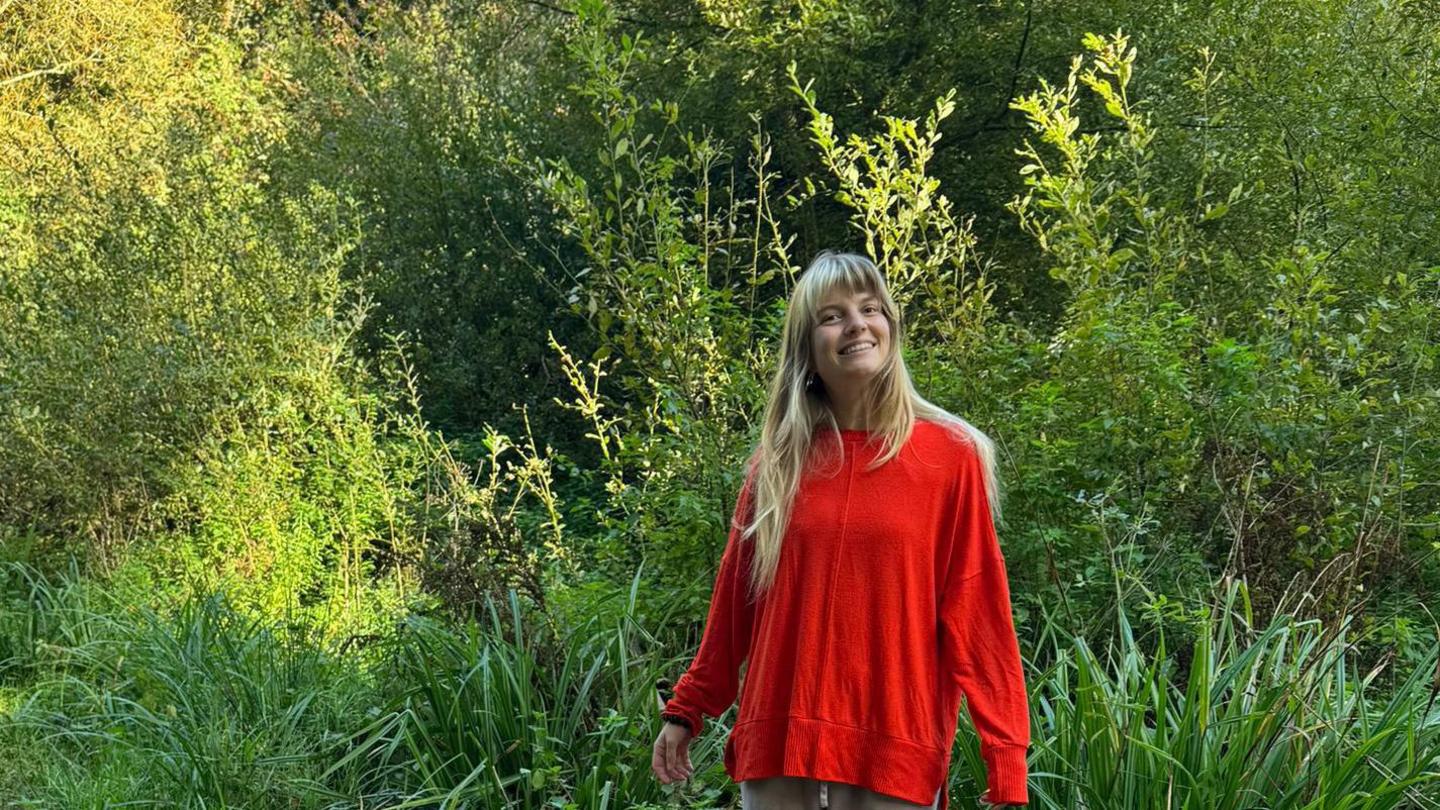 A woman wearing a red jumper standing in a woodland setting. Green trees are behind her. She is smiling at the camera.