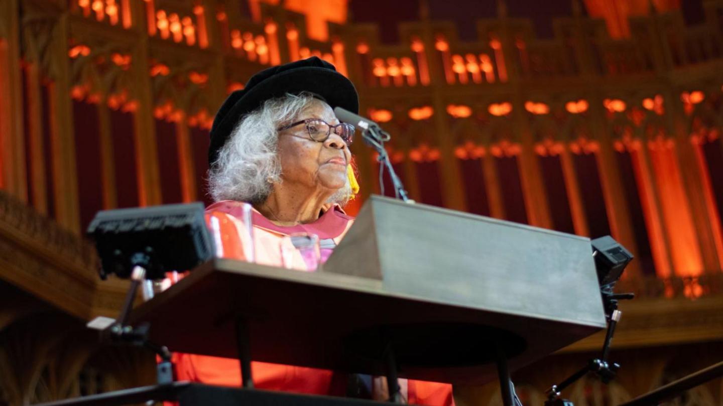 Barbara Dettering standing behind a black podium giving a speech. She is wearing her red graduation robes and her black graduation cap