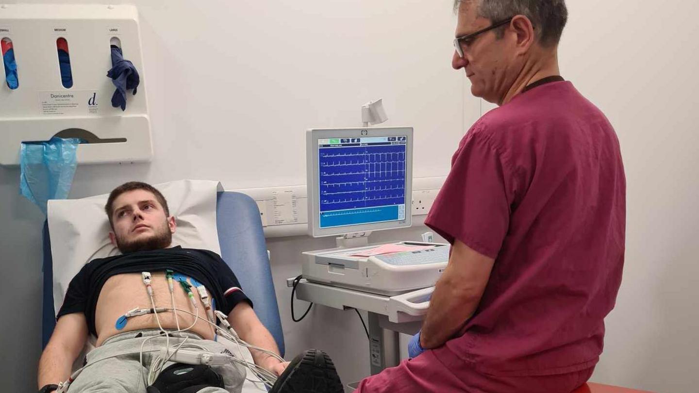 Louis lying on a hospital bed with wires attached next to his desk, while a member of hospital staff in scrubs watches