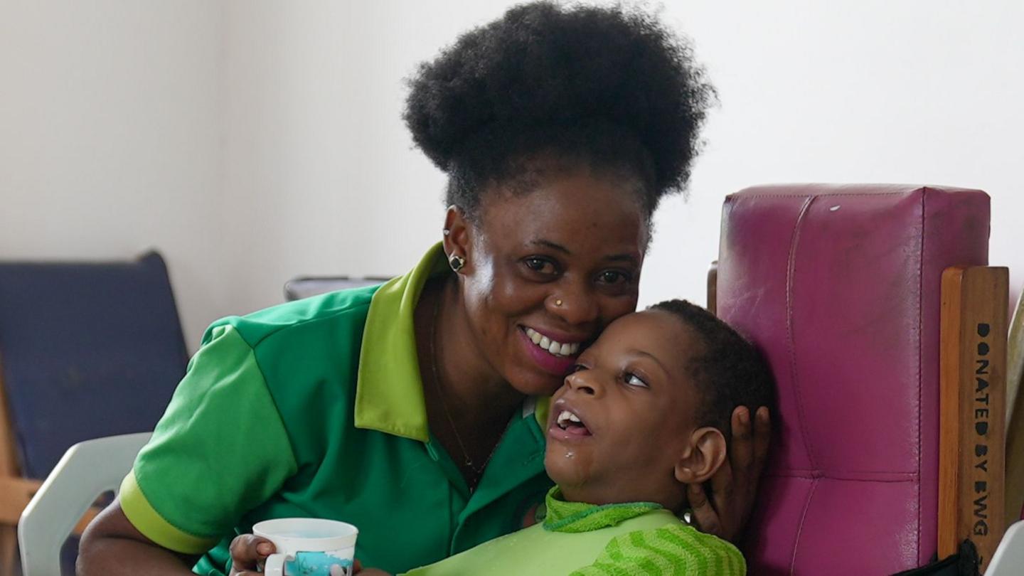 A woman wearing green holds a mug with one hand and the back of a child's head with the ther. The child is also wearing green.