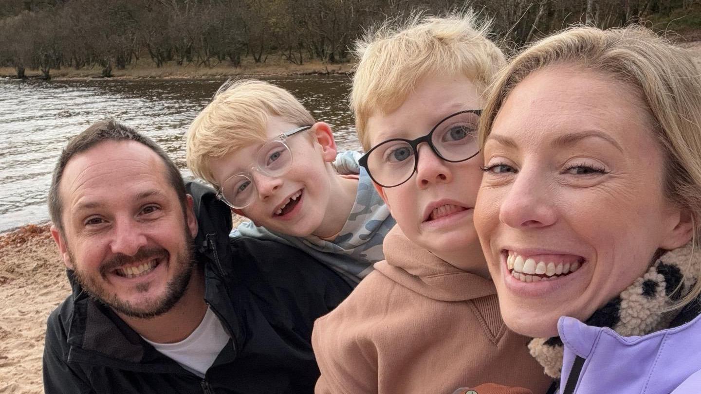 Daisy Woods, pictured right, smiles in a selfie with her husband Andy (left), son Oscar (second left) and son Sonny (second right). They are on a beach with trees in the background and all are wearing coats and fleeces.